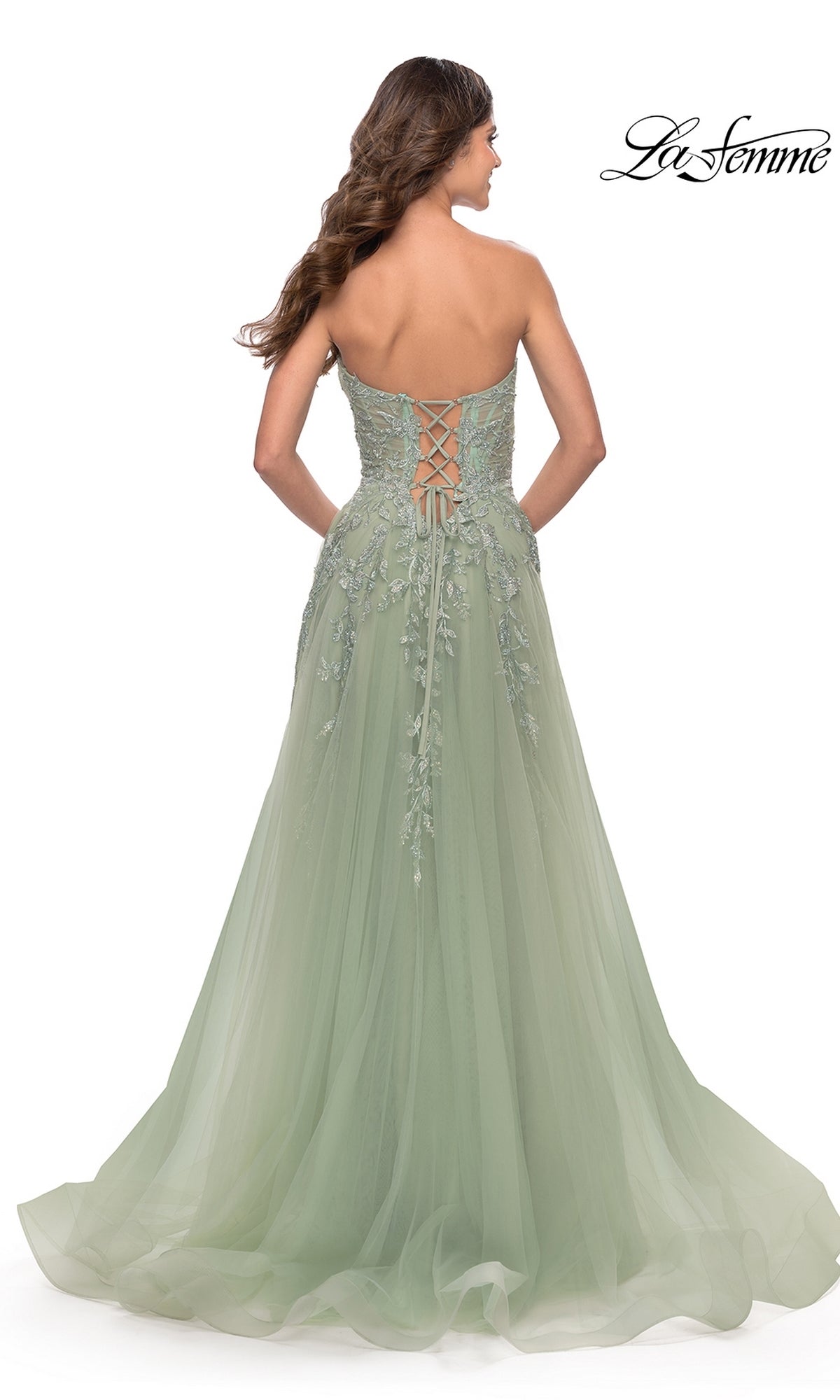 A woman stands with her back to the camera, wearing the La Femme Strapless Sweetheart Long Prom Dress 31363 in sage green, featuring a lace-up back and floral embroidery. The